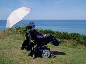 Woman in wheelchair, seen from behind, sat at the edge of a cliff looking out to sea. A parasol shades her from the sun