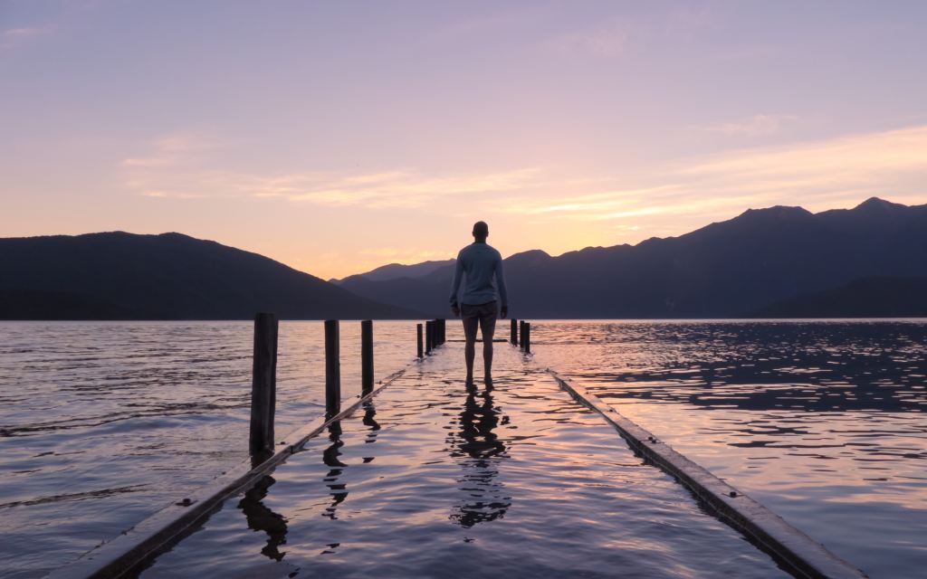 A person stands at the edge of a lake, looking out at the vast body of water. Their feet are submerged by water.