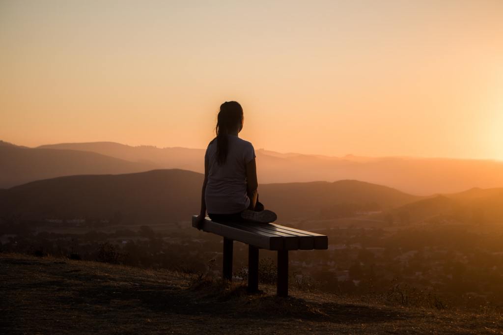 A silhouetted figure sits on a bench, looking out over hills.  The sun is setting over the scene.