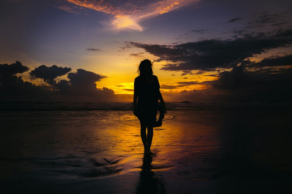 A silhouetted woman stands at the edge of the sea. The sun is setting and the sky is a mixture of dark clouds and golden light. The sea in front of her is reflecting the sunlight.